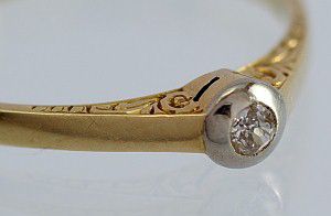 Prsten ze zlata ryzosti 585/1000 (14 karátů), briliant velikosti 0,05 ct, práce ze 30. let 20. stol. s pečlivě provedenou rytinou u hlavy prstenu. Samotný kámen zasazen v bílém zlatě.